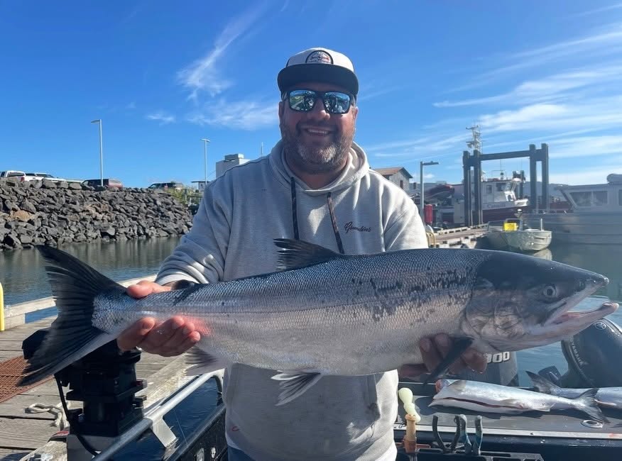 A man wearing a baseball cap and a grey hoodie smiles while holding a large fish